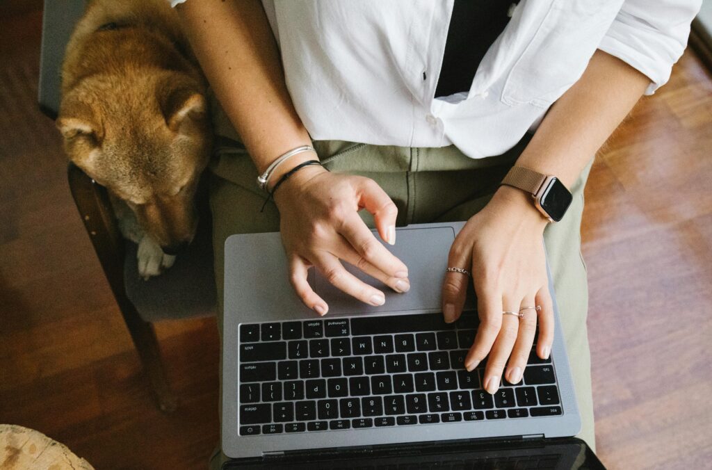 Woman wearing an apple watch working on a laptop, sitting next to her dog.